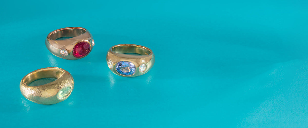 Image of group of yellow gold jewels set with emeralds and sapphires on a turquoise background